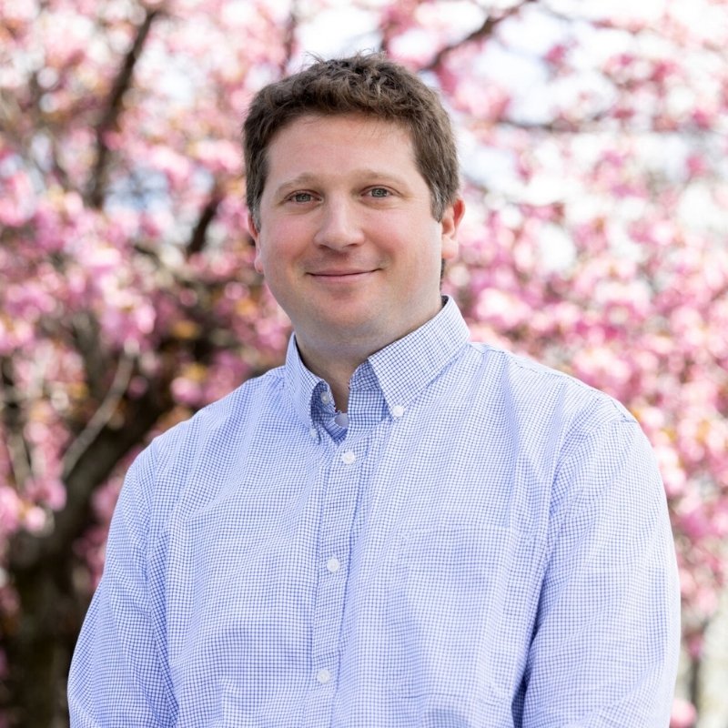 Dr. Peter Kleckner, audiologist, smiling in a light blue checked shirt in front of a blossoming pink tree